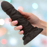 A hand with manicured nails holds the CalExotics Monster Dong The Mummy, a large black silicone dildo with textured surface, suction base, and attached vibrating bullet, against a blurred colorful background.